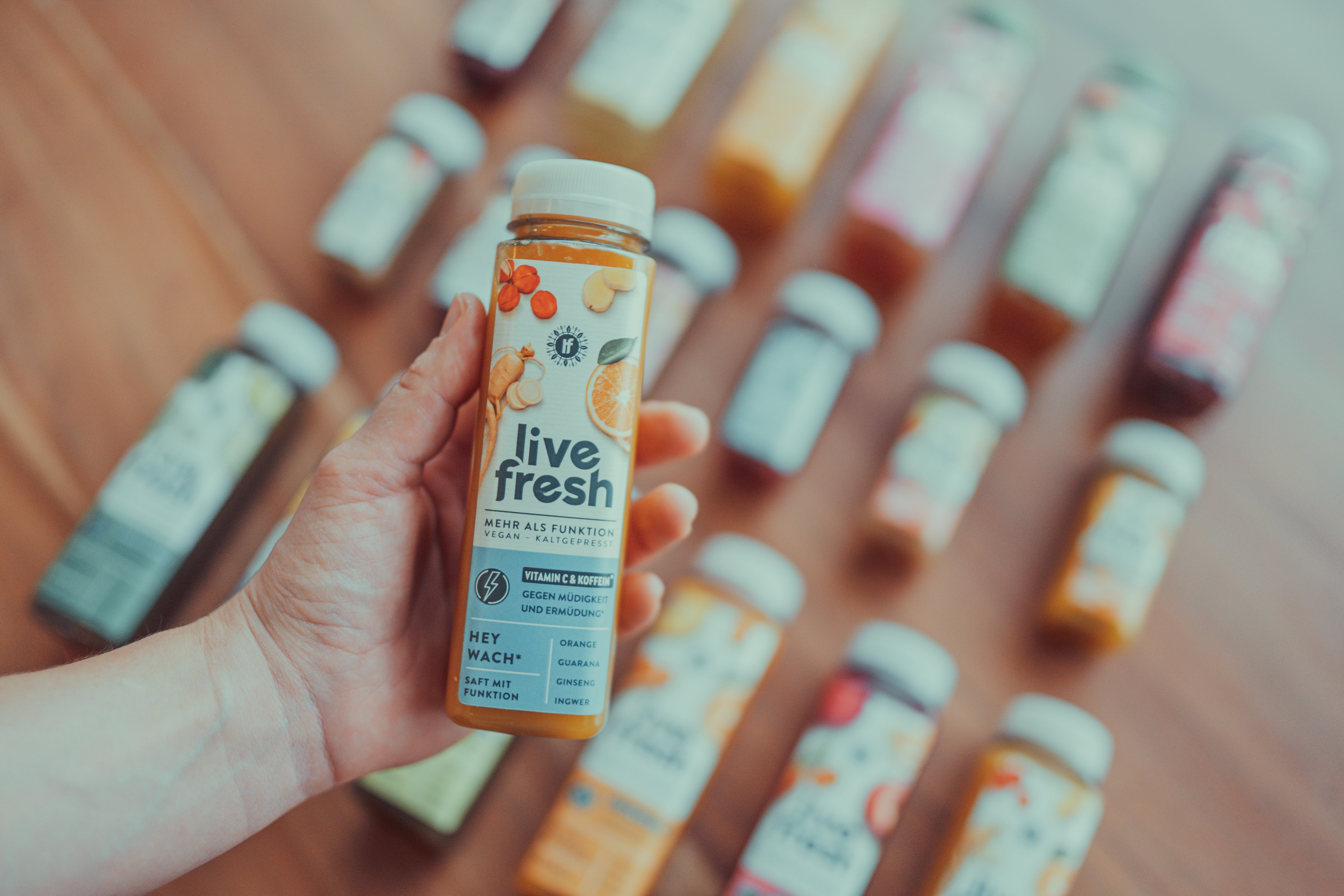 Eine Hand, die eine Flasche „Live Fresh“ Saft mit der Aufschrift „Hey Wach!“ hält. Die Flasche hebt hervor, dass der Saft mit Vitamin C und Koffein gegen Müdigkeit wirkt. Im Hintergrund sind weitere „Live Fresh“-Säfte in verschiedenen Sorten unscharf auf einem Holztisch verteilt. Die Flaschen sind in unterschiedlichen Farben gehalten, die die Vielfalt der angebotenen Geschmacksrichtungen und Funktionen betonen.