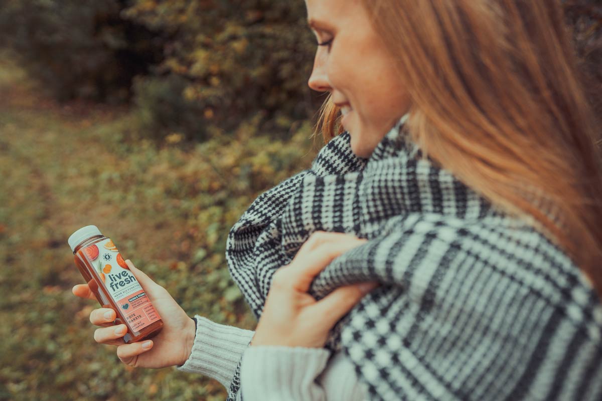 Eine Frau im Freien, die eine Flasche „Live Fresh Rote Rakete“ Saft in der Hand hält. Sie ist in einen schwarz-weiß karierten Schal gehüllt, während sie lächelnd die Saftflasche betrachtet. Die herbstliche Umgebung mit Bäumen und Laub verleiht dem Bild eine gemütliche Atmosphäre.