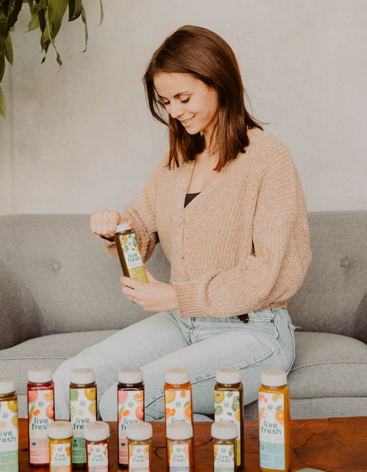 Junge Frau, die auf einem grauen Sofa sitzt und eine „Live Fresh“-Saftflasche öffnet. Vor ihr auf dem Tisch stehen mehrere Flaschen verschiedener „Live Fresh“-Säfte in unterschiedlichen Geschmacksrichtungen. Die Frau lächelt und wirkt entspannt, während sie den Saft vorbereitet. Die Szene vermittelt eine ruhige und gesunde Atmosphäre, mit Fokus auf die frischen, farbenfrohen Säfte.